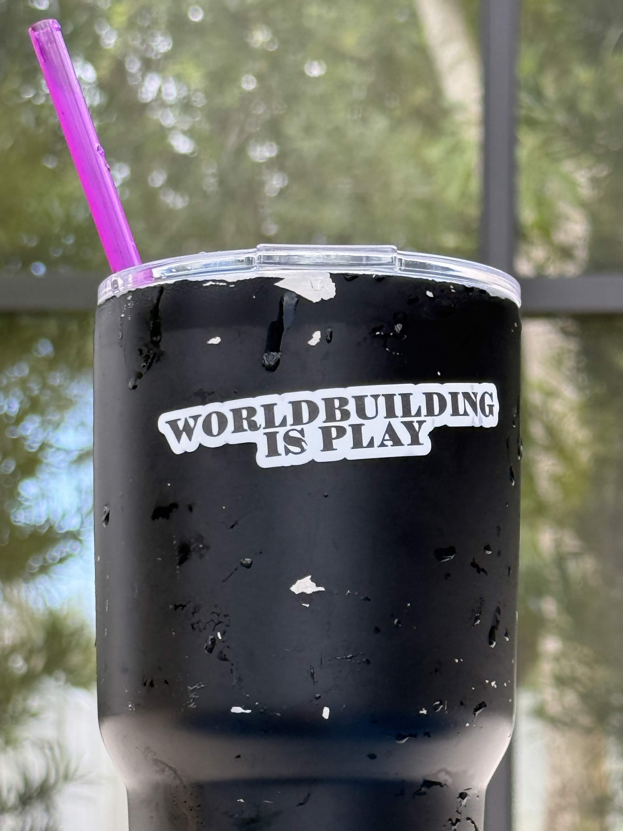 image Black tumbler with peeling paint and white text reading 'WORLDBUILDING IS PLAY' with a purple straw